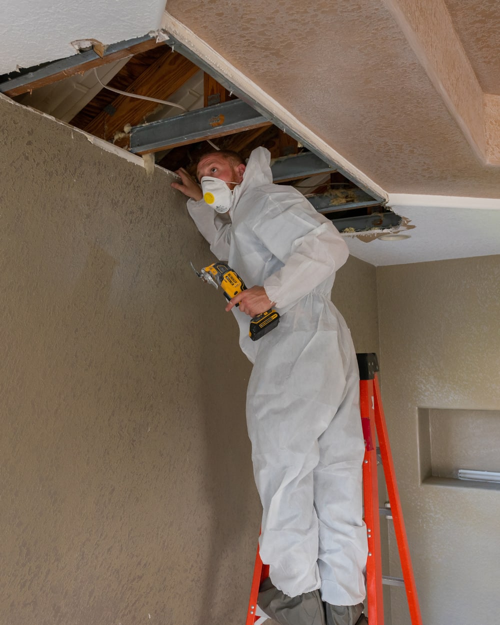 Restoration technician in protective coveralls opening a water-damaged ceiling to reach the wet cavity above